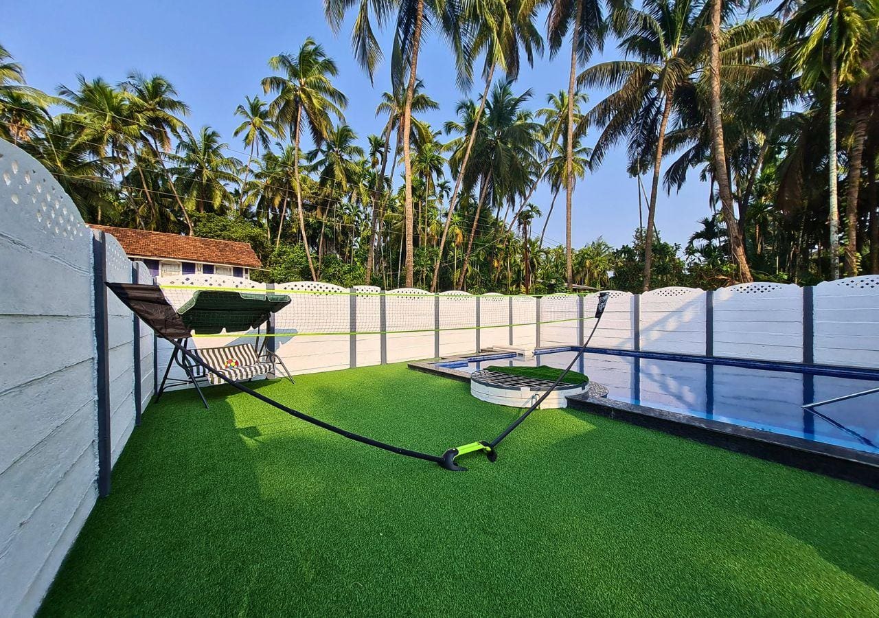 lush green lawn with black & neon badminton net, a green swing with white stripes, a swimming pool, well and white fence.